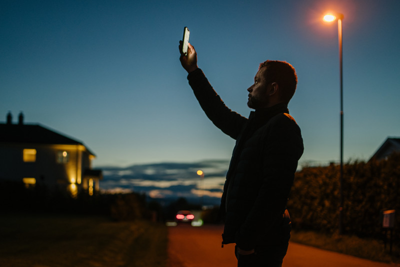 Mann utendørs på kveldstid som holder en mobiltelefon opp i luften